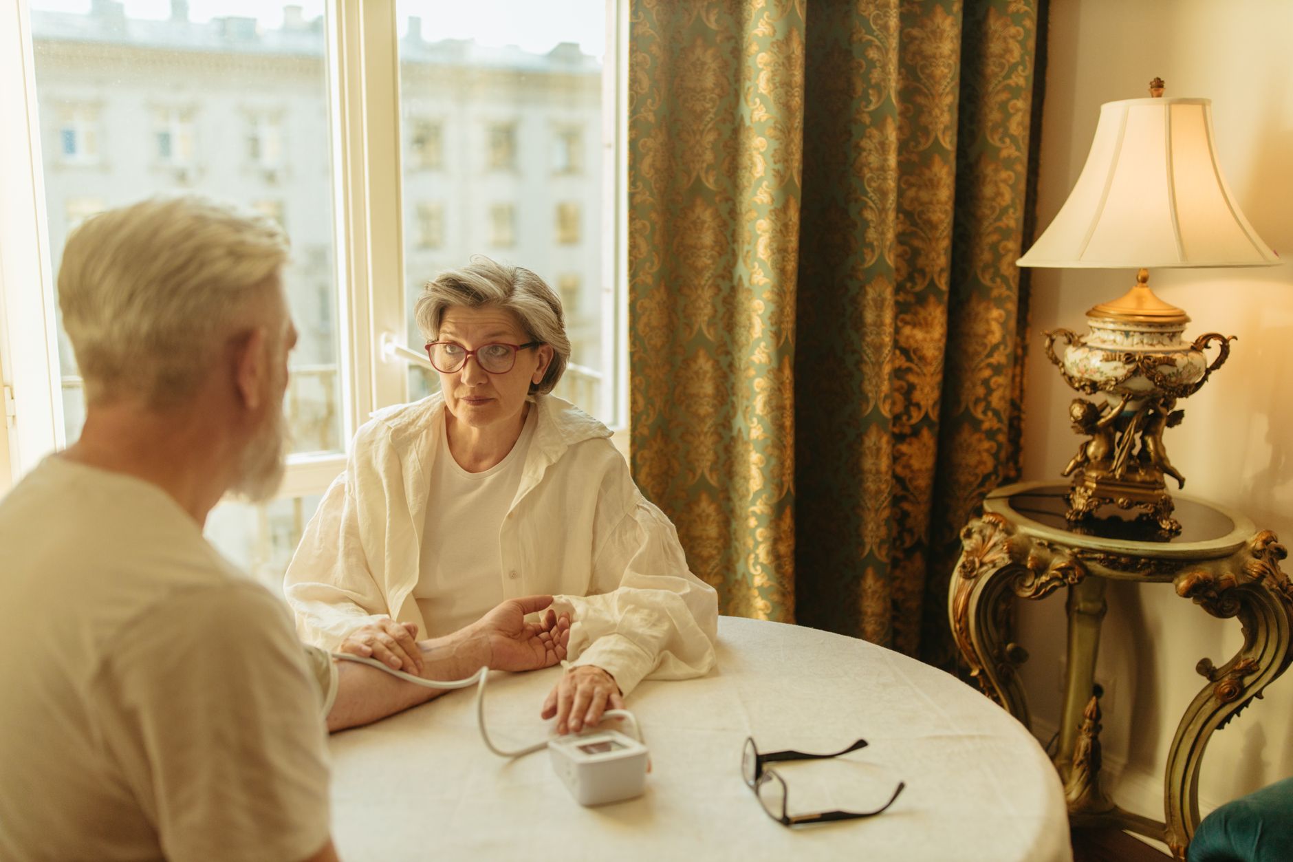 Senior woman checks man's blood pressure at home. Warm light, caring interaction.