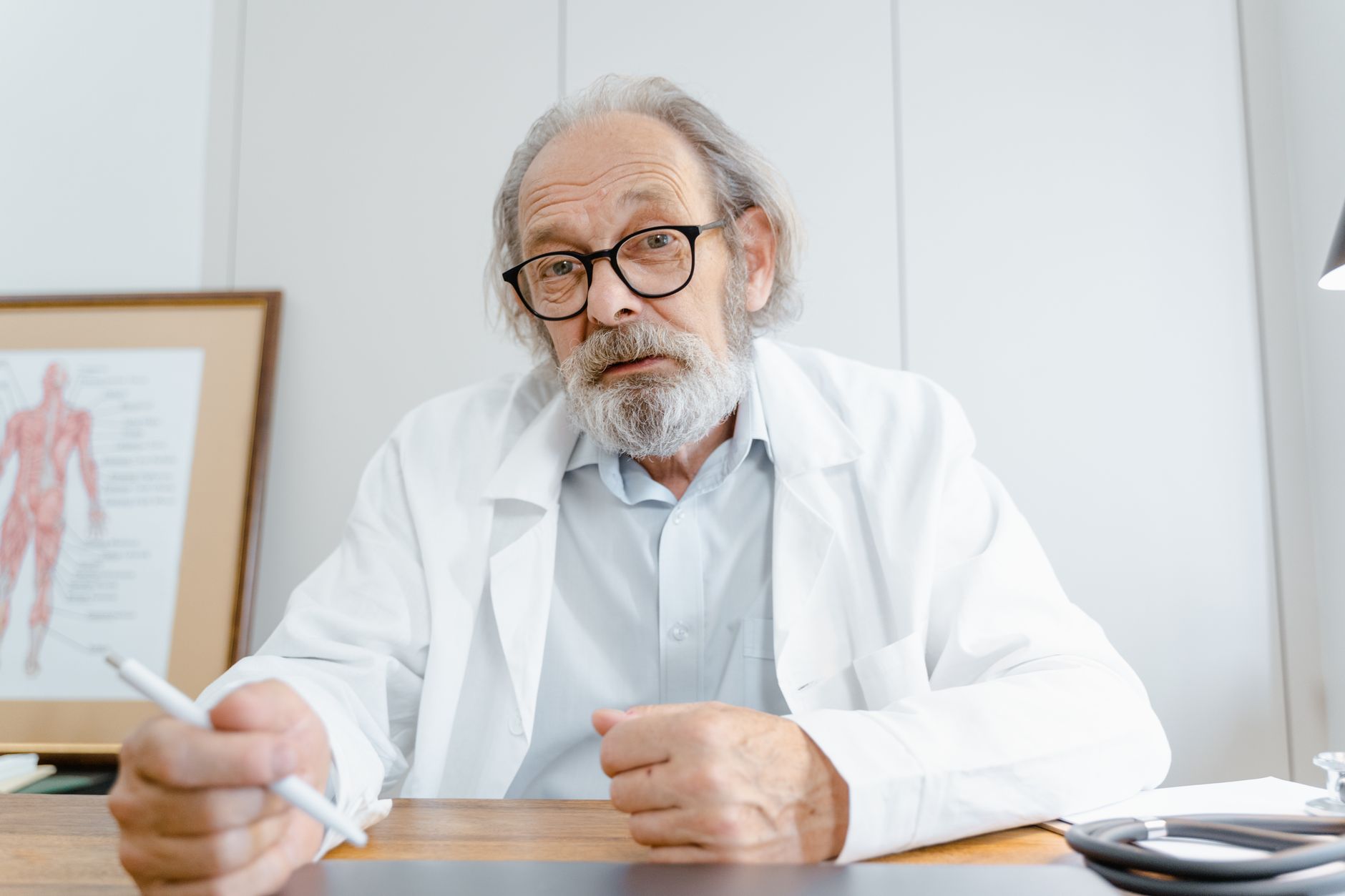 Elderly doctor with glasses and beard in office setting, holding a pen, emphasizing telemedicine professionalism.