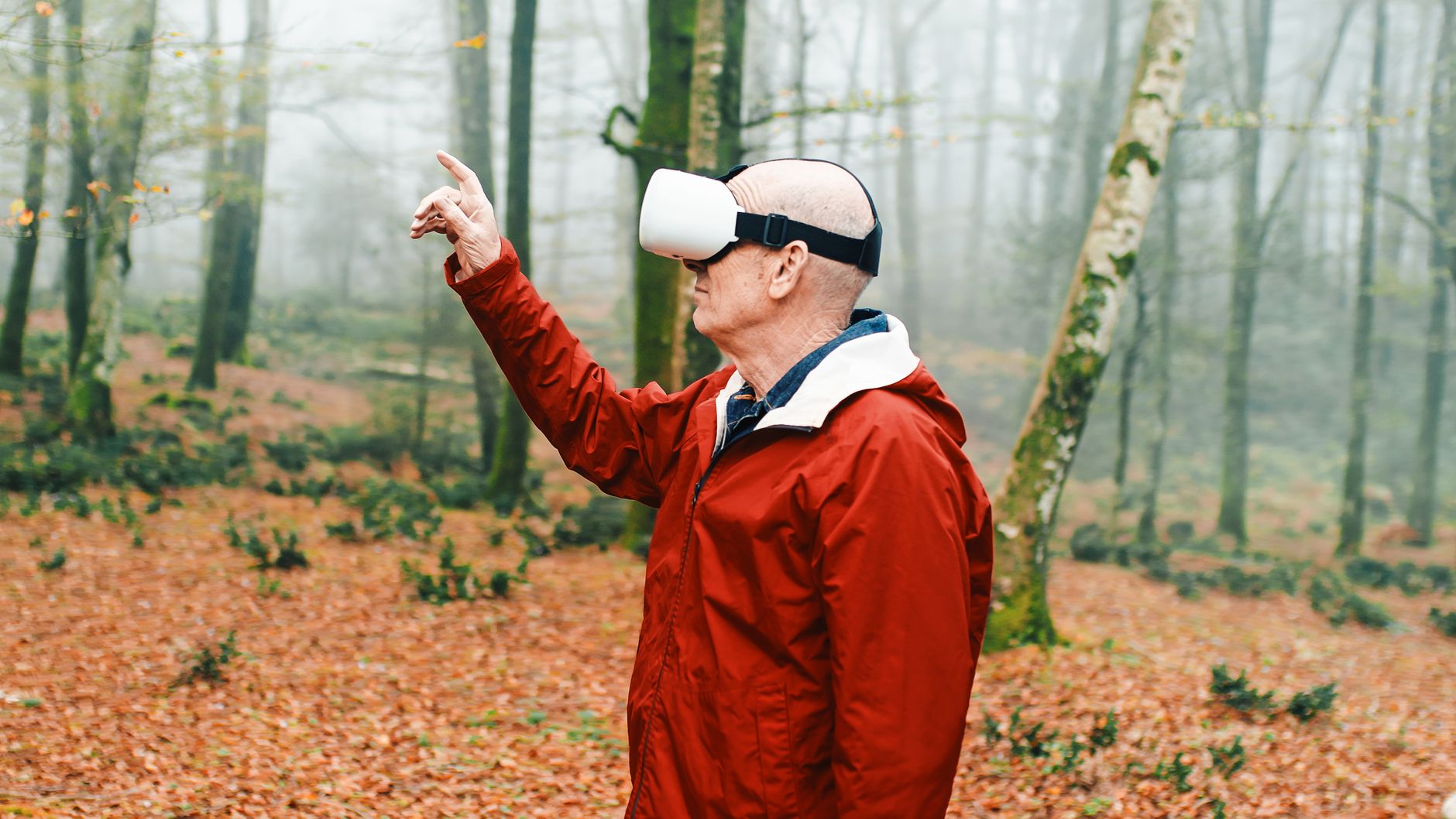 An elderly man in a red jacket interacts with virtual reality in an autumn forest setting.