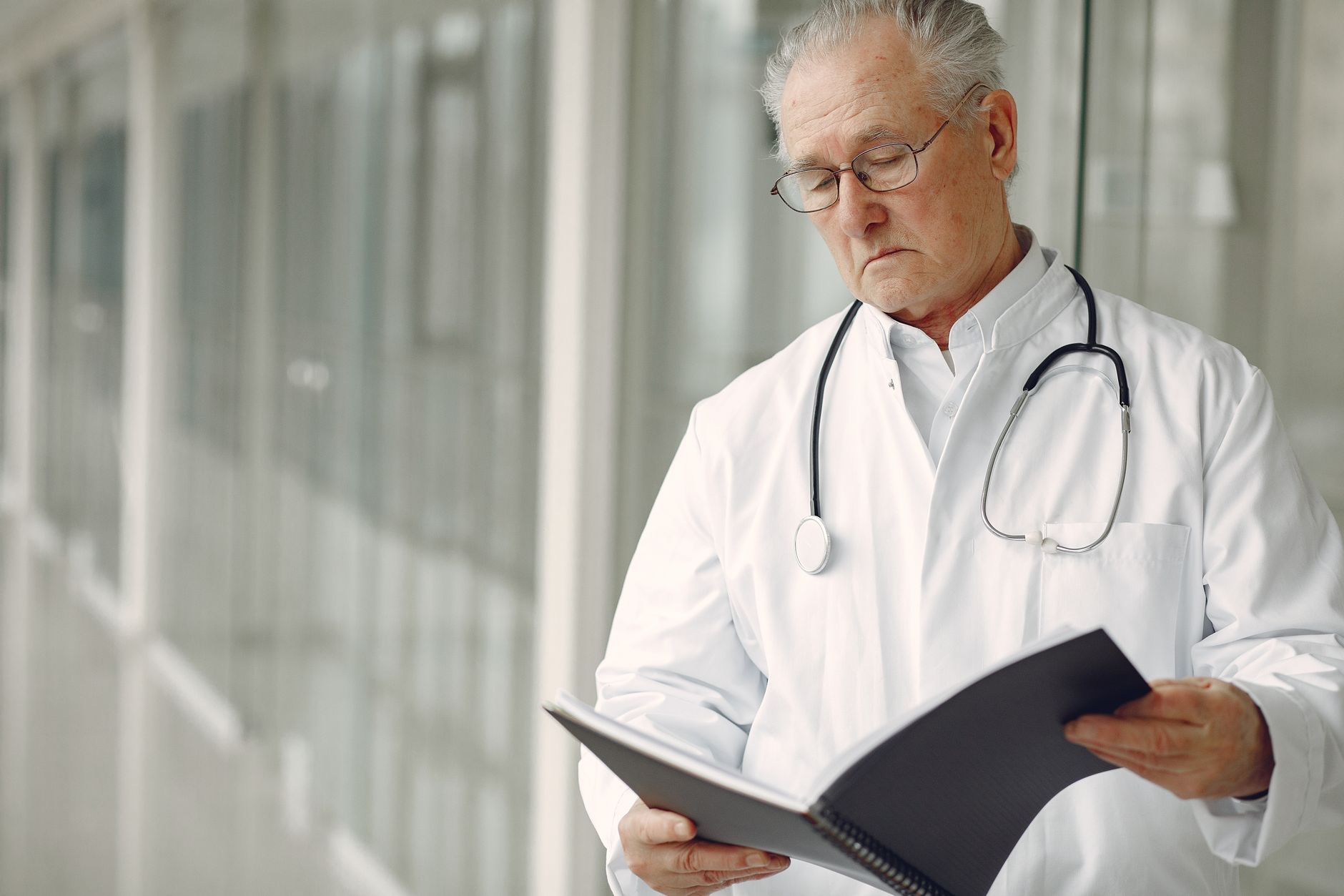 Crop concentrated professional senior male physician in white robe with stethoscope standing in modern hospital hallway and reading medical case records in daylight