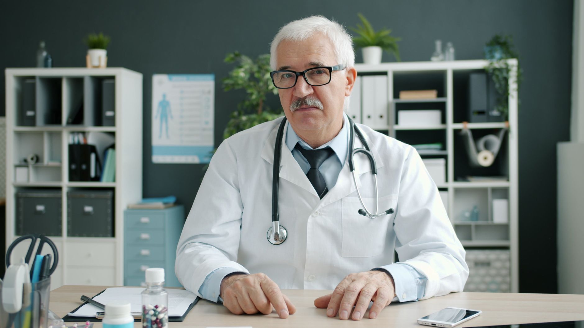 Senior doctor in an office setting, with medical equipment and files in the background.