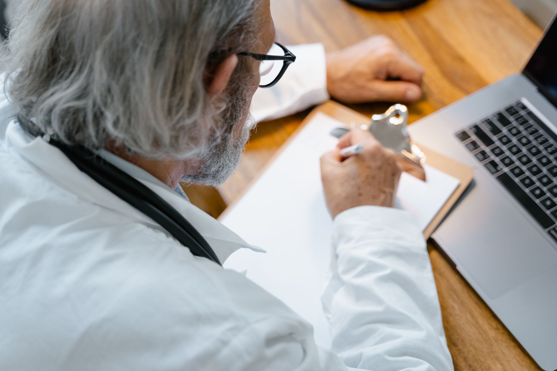 An elderly doctor in a white coat writing notes on a clipboard at a desk with a laptop.